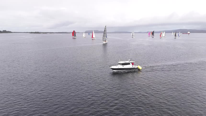 Aerial view of a support motorboat cruising alongside a fleet of sailboats with colorful spinnakers during a regatta on a cloudy day