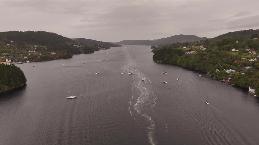 A scenic aerial view of sailboats and motorboats navigating a long fjord in Norway under a cloudy, overcast sky