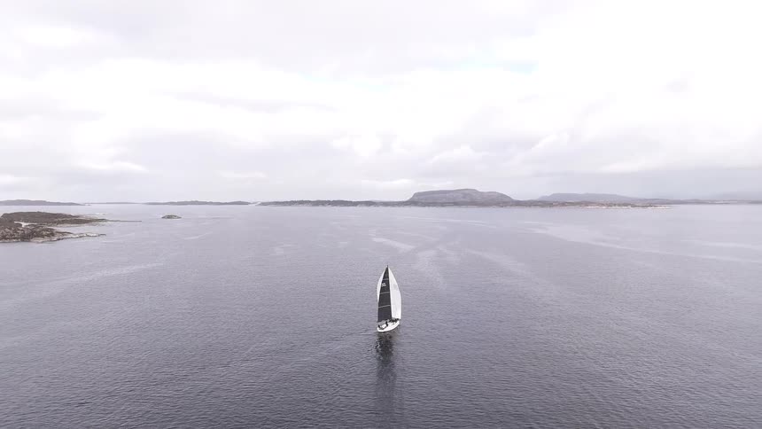 A drone pulls back to reveal a lone sailboat navigating a vast, tranquil sea along a rocky coastline under a cloudy sky