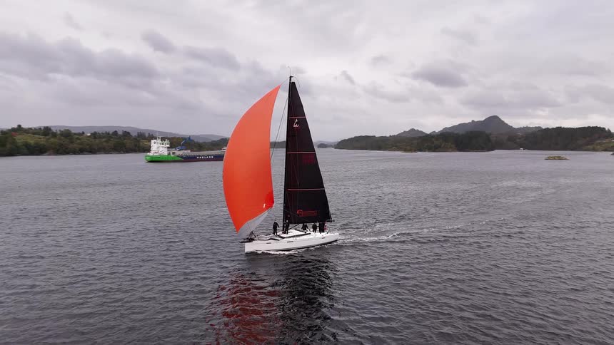 An aerial view follows a racing sailboat with a vibrant orange spinnaker as it gracefully sails across a fjord during a regatta