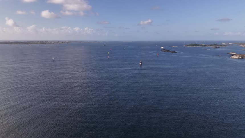 A serene, static aerial view shows a sailboat race on the vast blue ocean surrounded by a rocky archipelago under a partly cloudy sky