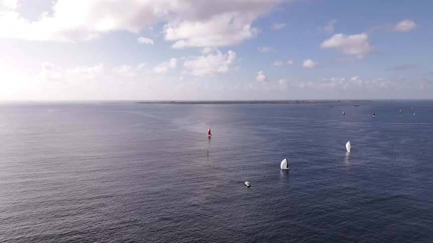 A wide, static aerial view shows sailboats scattered across the vast, calm ocean under a bright, partly cloudy sky