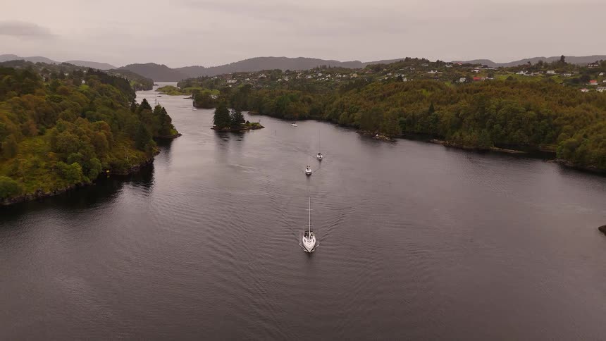 An aerial view shows a fleet of sailboats cruising in a line through a tranquil fjord on an overcast day