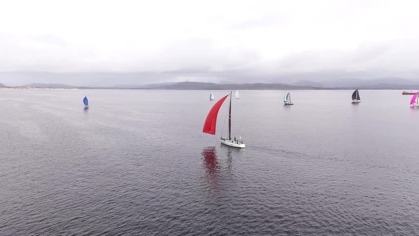An aerial view captures a fleet of sailboats with colorful spinnakers gliding across the open sea during a regatta on a cloudy day