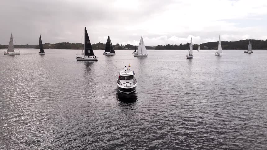 A spectator motorboat watches a fleet of sailboats compete in a regatta on a calm sea under an overcast sky