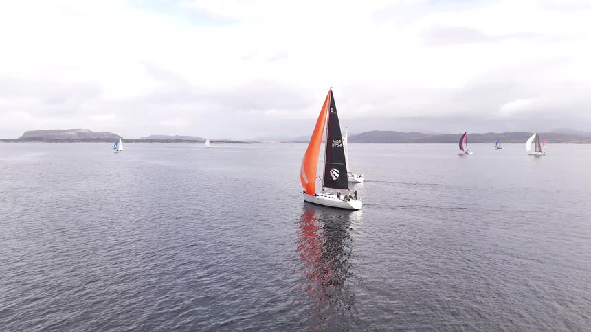 A fleet of sailboats with colorful spinnakers participate in a regatta on a calm sea under a cloudy sky