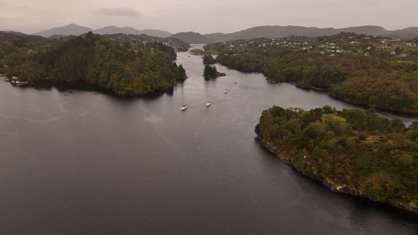 An aerial view captures several sailboats as they peacefully navigate a picturesque waterway between forested islands on a calm, overcast day