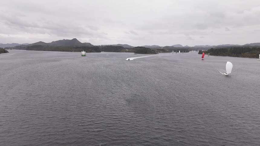 A wide aerial view shows a sailing regatta and a motorboat cruising along a scenic channel on a cloudy day