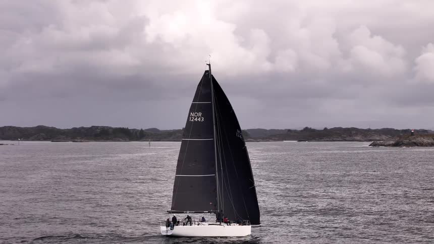 An aerial pullback shot reveals a sleek racing sailboat with a large black sail cruising through moody waters past rocky islands on an overcast day