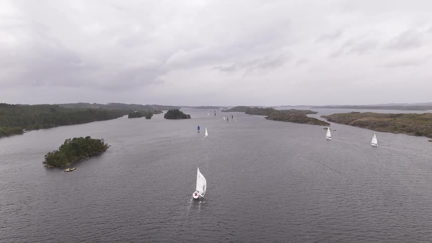 An aerial drone shot slowly rises to reveal a regatta of sailboats navigating through a scenic channel surrounded by islands on an overcast day