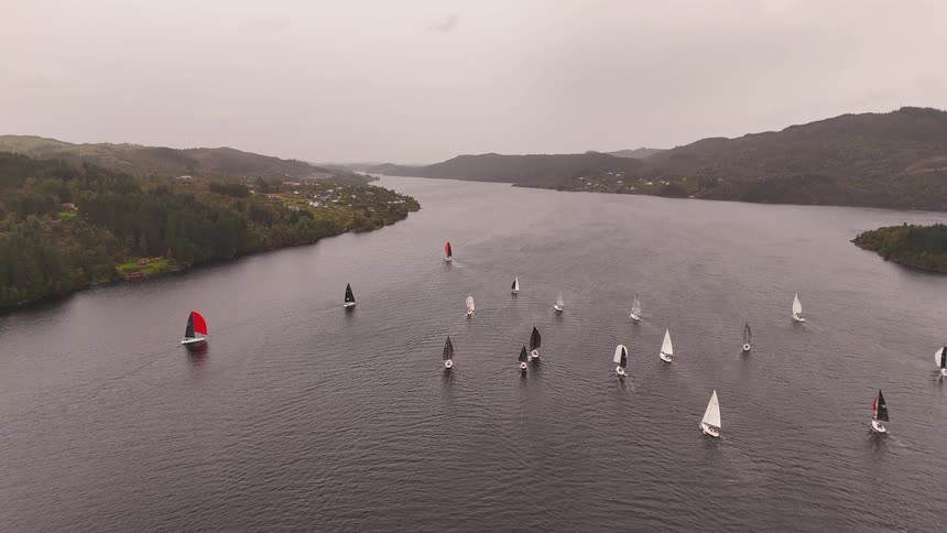 An aerial drone shot captures a scenic sailboat regatta on a wide loch surrounded by green hills on a cloudy day