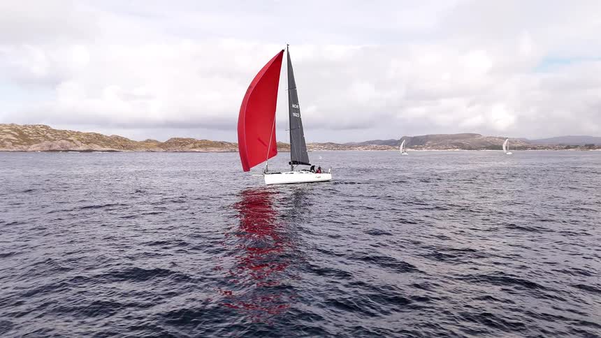 A sleek, modern sailboat with a striking red spinnaker glides across the water during a regatta on a cloudy day