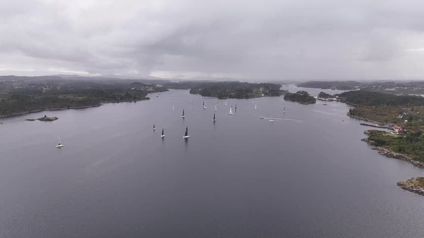 A wide, static aerial shot captures a sailing regatta on a vast fjord surrounded by forested islands on an overcast day