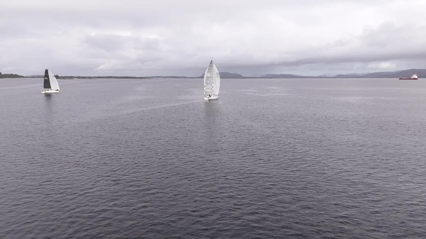 Aerial footage follows a fleet of sailboats competing in a regatta on a vast, open sea under a grey, cloudy sky