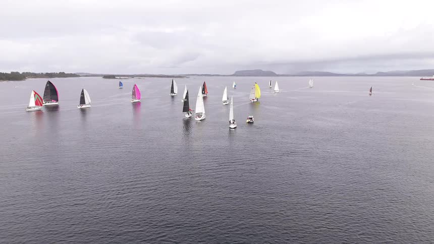 Aerial view of a regatta with numerous sailboats featuring colorful spinnakers racing across the water on a cloudy day