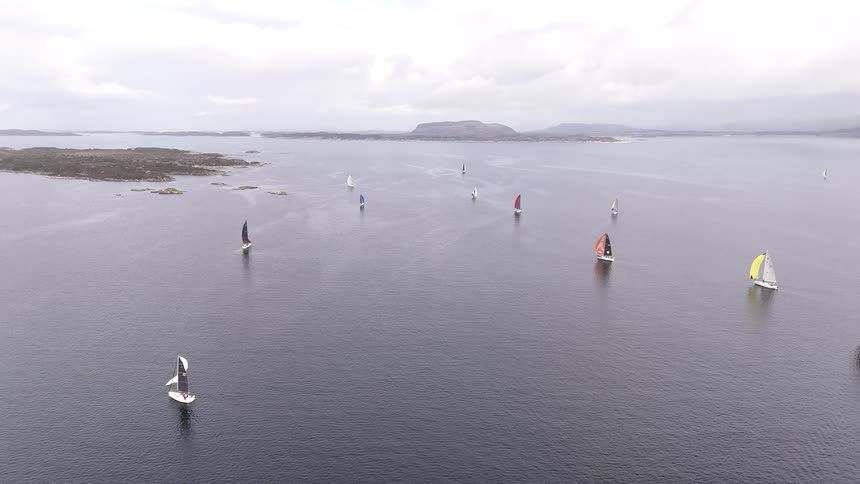 A serene aerial view shows a fleet of sailboats with vibrant, colorful sails participating in a regatta on calm waters under a cloudy sky