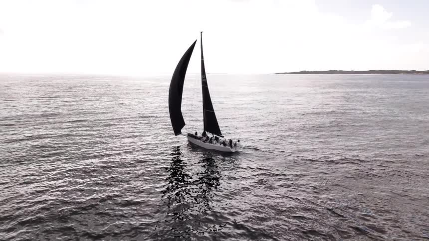 An aerial view follows a sailboat with a large black sail gliding across the open sea, transitioning from color to dramatic black and white