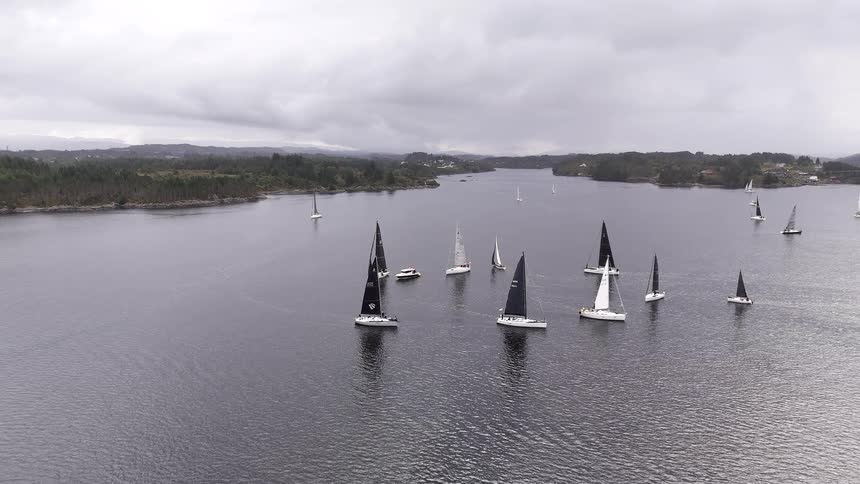 A high-angle aerial shot slowly pulls back to reveal a fleet of sailboats racing on a vast fjord on a cloudy day