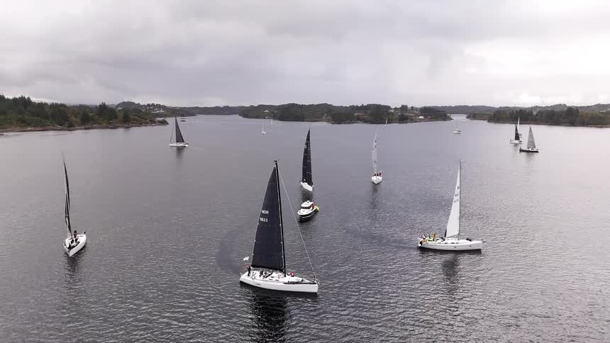 An aerial view shows a fleet of sailboats with black and white sails competing in a regatta on a calm fjord under a cloudy sky