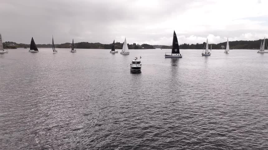A spectator motorboat navigates through a fleet of sailboats participating in a regatta on a cloudy day