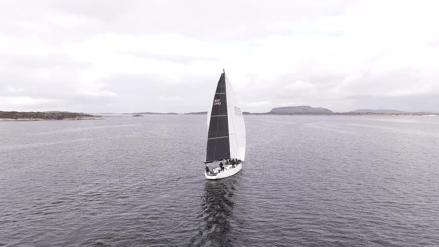 An aerial shot follows a modern sailboat with large black and white sails as it glides across the calm water on an overcast day