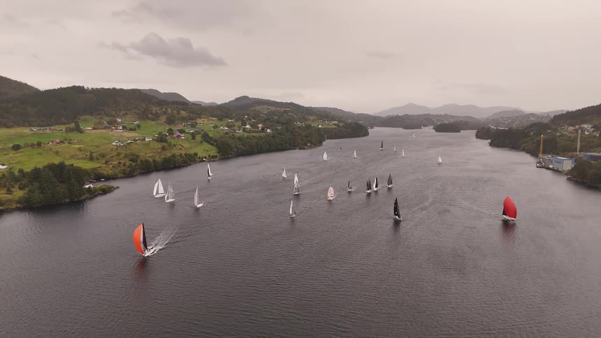 An expansive aerial view captures a fleet of sailboats with colorful spinnakers participating in a regatta on a wide river on a cloudy day