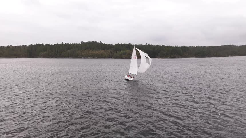 An aerial shot follows a sailboat with a large white spinnaker as it sails across a body of water on a cloudy day