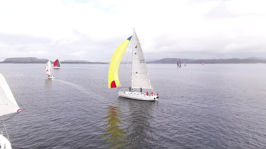 Aerial footage captures a fleet of sailboats with colorful sails competing in a regatta on a calm sea under a cloudy sky
