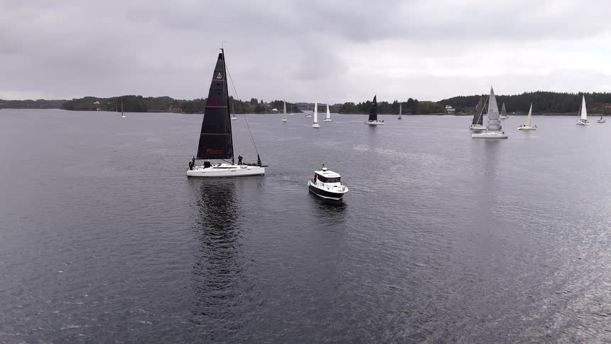 A fleet of sailboats and a motorboat gather on a calm body of water for a regatta on a gray, overcast day