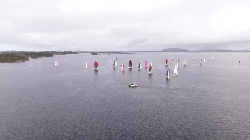 Aerial view flying away from a large group of sailboats with colorful sails competing in a regatta on a vast, calm sea under an overcast sky