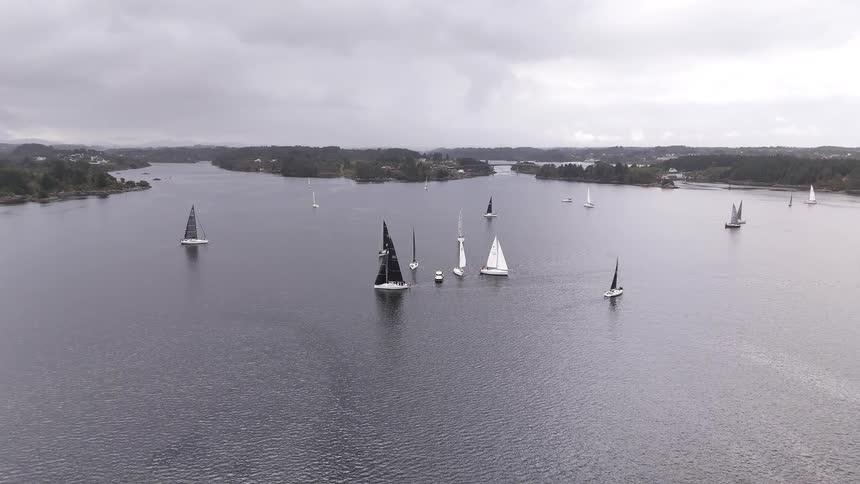 A fleet of sailboats races across a wide, tranquil channel between green islands under a cloudy sky