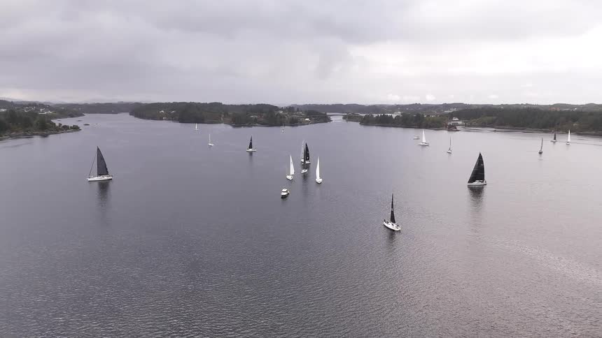 A serene aerial view pulls back to reveal a fleet of sailboats gliding across a tranquil bay during a regatta on a cloudy day