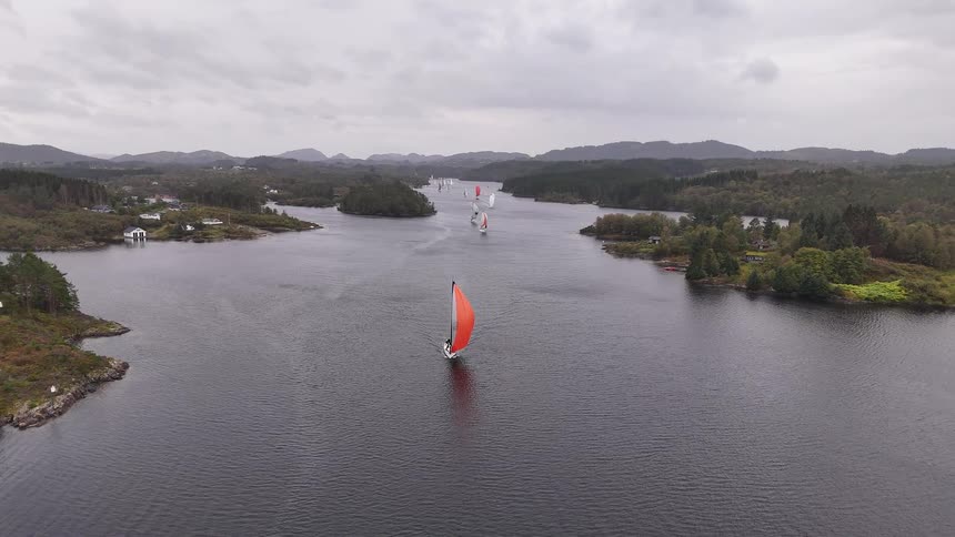 Aerial view of a sailboat with a striking red spinnaker leading a regatta through a scenic fjord on an overcast day