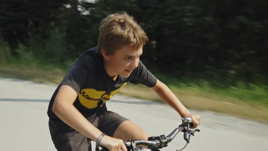 A determined young boy rides his bicycle quickly down a tree-lined road on a sunny day in slow motion