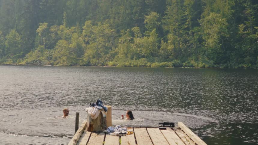 A young couple playfully jumps off a dock into a beautiful forested lake in slow motion