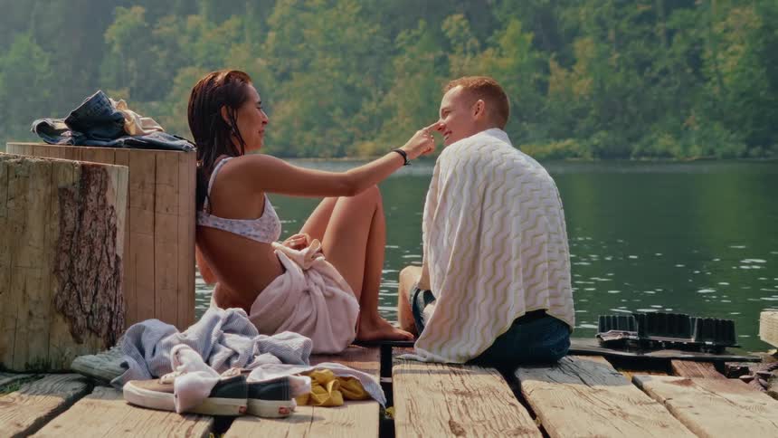 A happy young couple in towels flirts and talks on a rustic wooden dock by a beautiful, calm lake