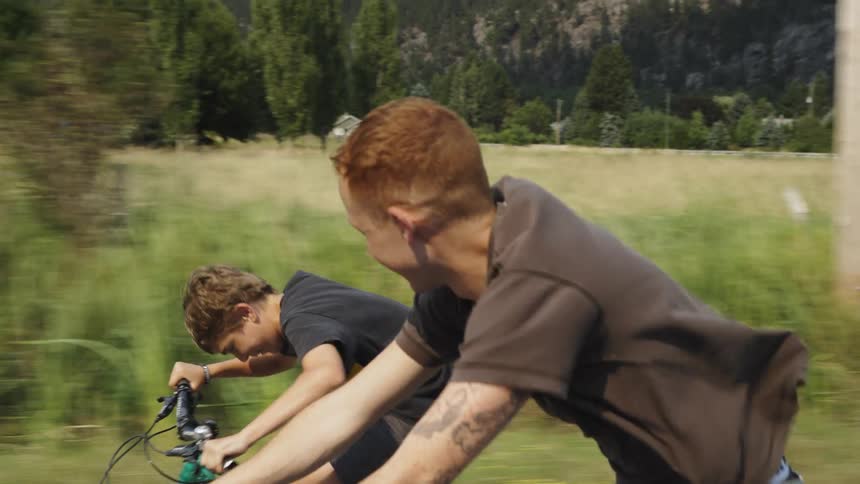 A slow-motion tracking shot captures two smiling young men enjoying a bike ride down a scenic country road