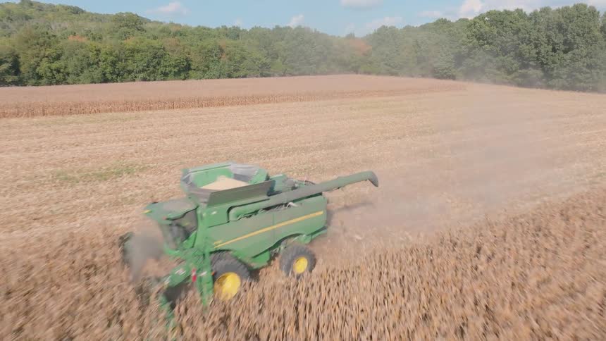 Aerial footage follows a large green combine harvester as it cuts through a vast, dusty field of soybeans during the fall harvest