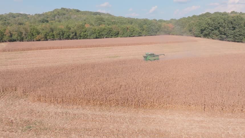 A wide aerial shot pushes in on a combine harvester as it gathers crops from a large field on a sunny day
