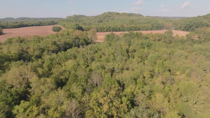 Aerial view slowly flying over a lush green forest with rolling hills and a farm field in the background on a sunny day