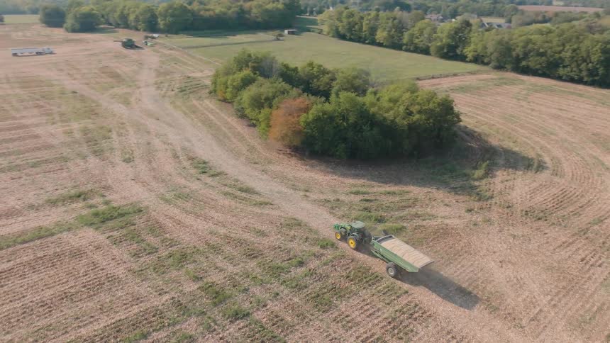 High angle aerial shot of a green tractor driving through a harvested field with a full grain cart