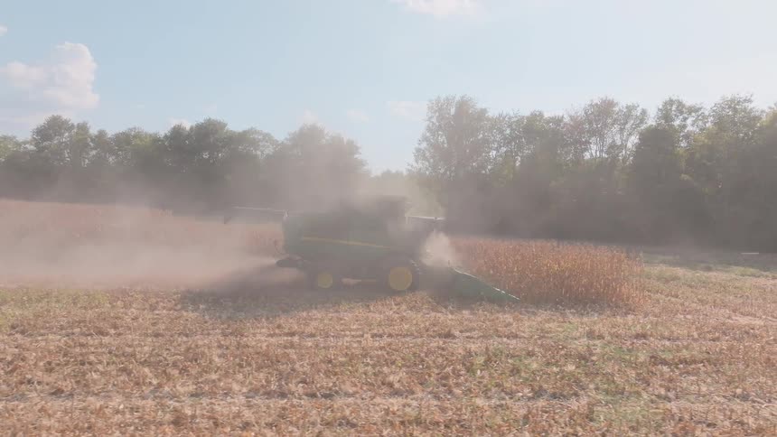 A green combine harvester creates a cloud of dust as it cuts through a dry field of crops in slow motion