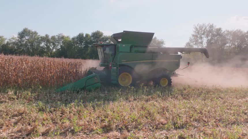 A slow-motion side view shows a green combine harvester kicking up dust as it harvests a dry cornfield on a sunny day