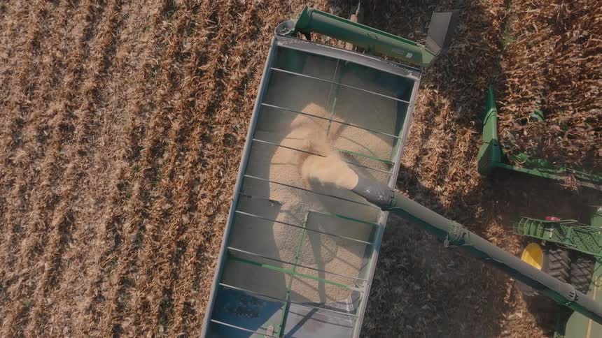 A top-down aerial view shows a combine harvester offloading corn into a grain cart while harvesting a field