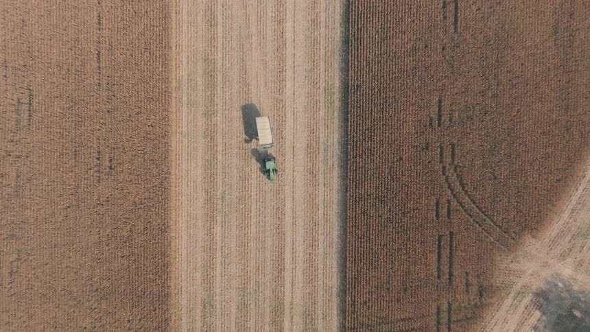 Top-down aerial footage of a green tractor with a trailer driving through a vast, harvested cornfield