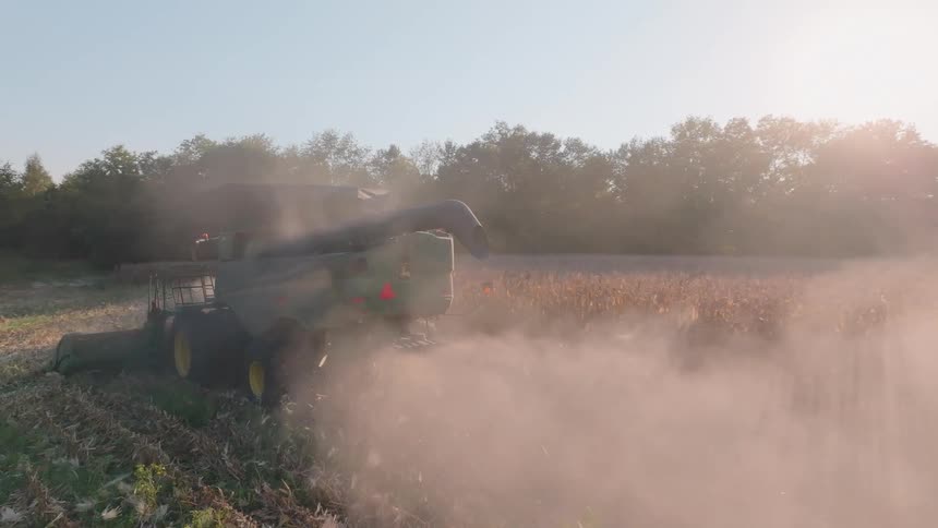 A combine harvester kicks up a thick cloud of dust as it harvests a crop field during a hazy golden hour