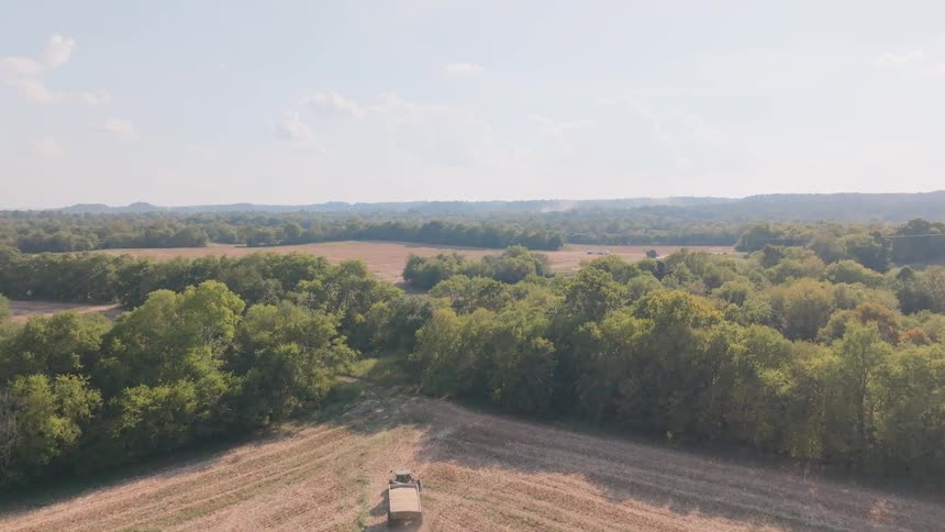 An aerial view follows a combine harvester working in a vast field before ascending to reveal the expansive rural landscape