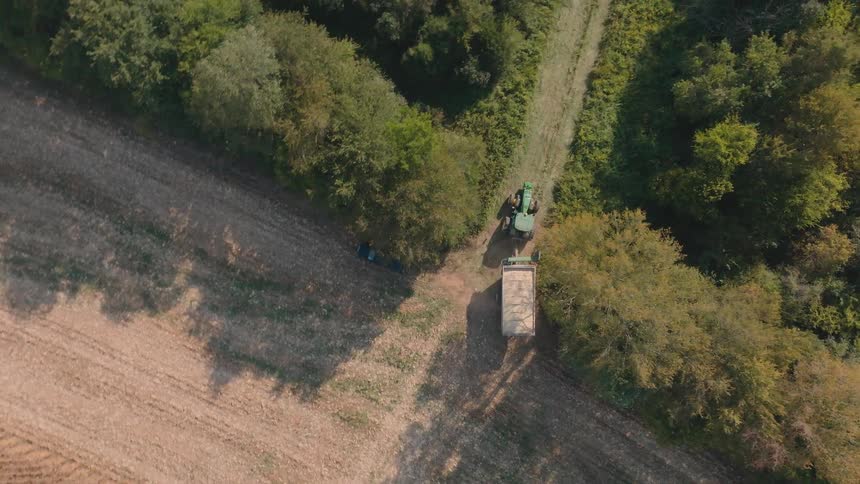 An aerial top-down view follows a green tractor pulling a full trailer from a harvested field onto a rural path leading into a dense forest