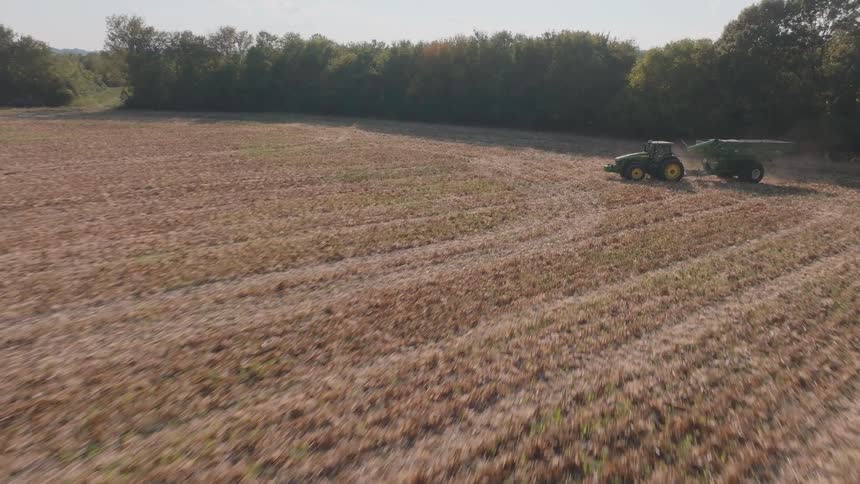 Aerial footage follows a combine harvester as it cuts through a dry cornfield, then flies over a waiting tractor and treeline