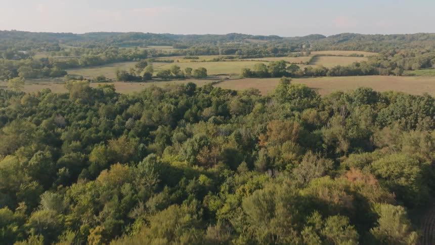 A smooth aerial reveal shot begins over a harvested crop field and rises to show a vast, green rural landscape